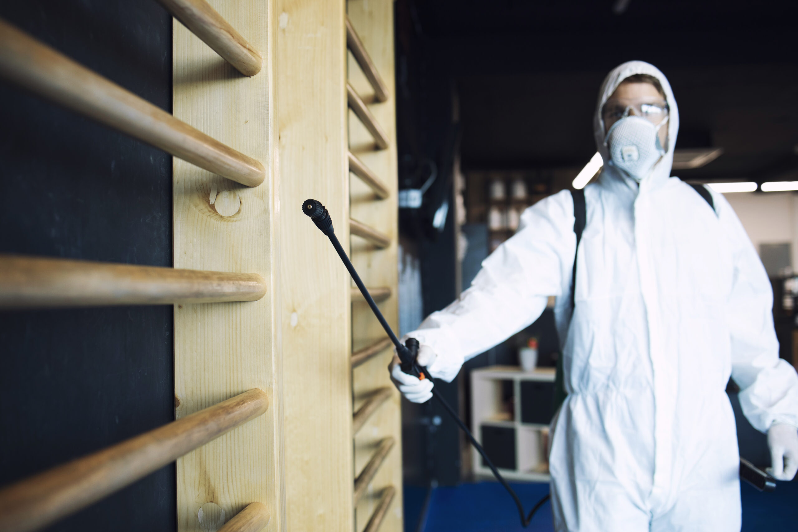 man in white protection suit disinfecting and fitness equipment and weights to stop spreading highly contagious corona virus.